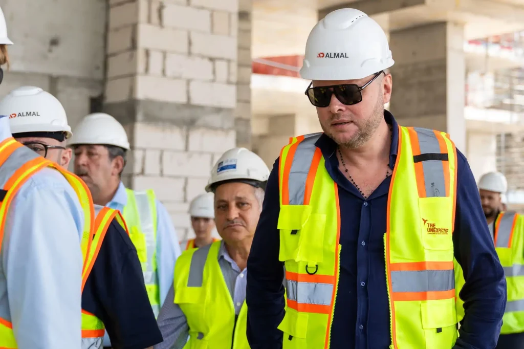 Group of construction workers in neon safety vests and white hard hats at a building site, with a man in sunglasses in the foreground.