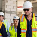 Group of construction workers in neon safety vests and white hard hats at a building site, with a man in sunglasses in the foreground.