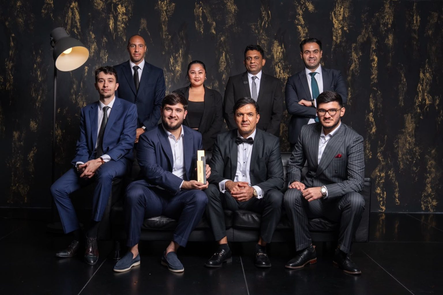 Group of well-dressed professionals posing in a studio; three men sit on a black sofa in front, one holds a trophy, others stand behind them against a dark, textured wall.